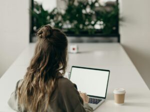 A woman with curly hair works on a laptop, facing a window with plants, symbolizing remote work.